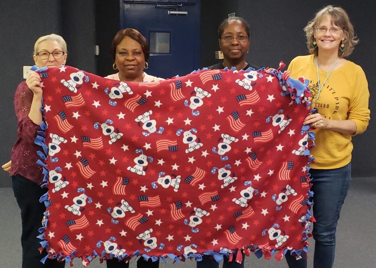 Four women hold up a large red blanket, featuring a pattern of a white dog and the American flag.