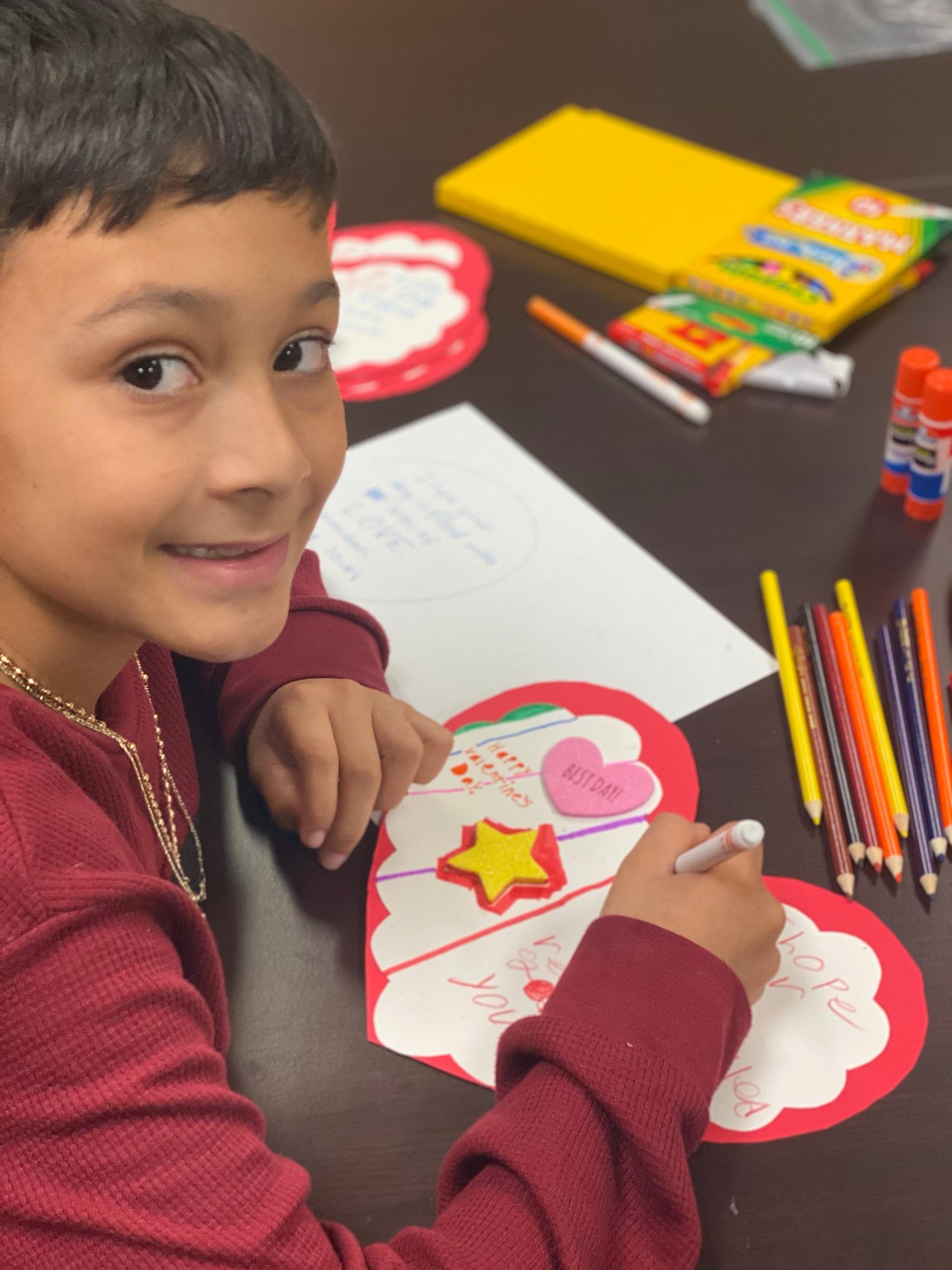 A boy with dark hair uses a marker to decorate heart-shaped, red card, surrounded by craft supplies.