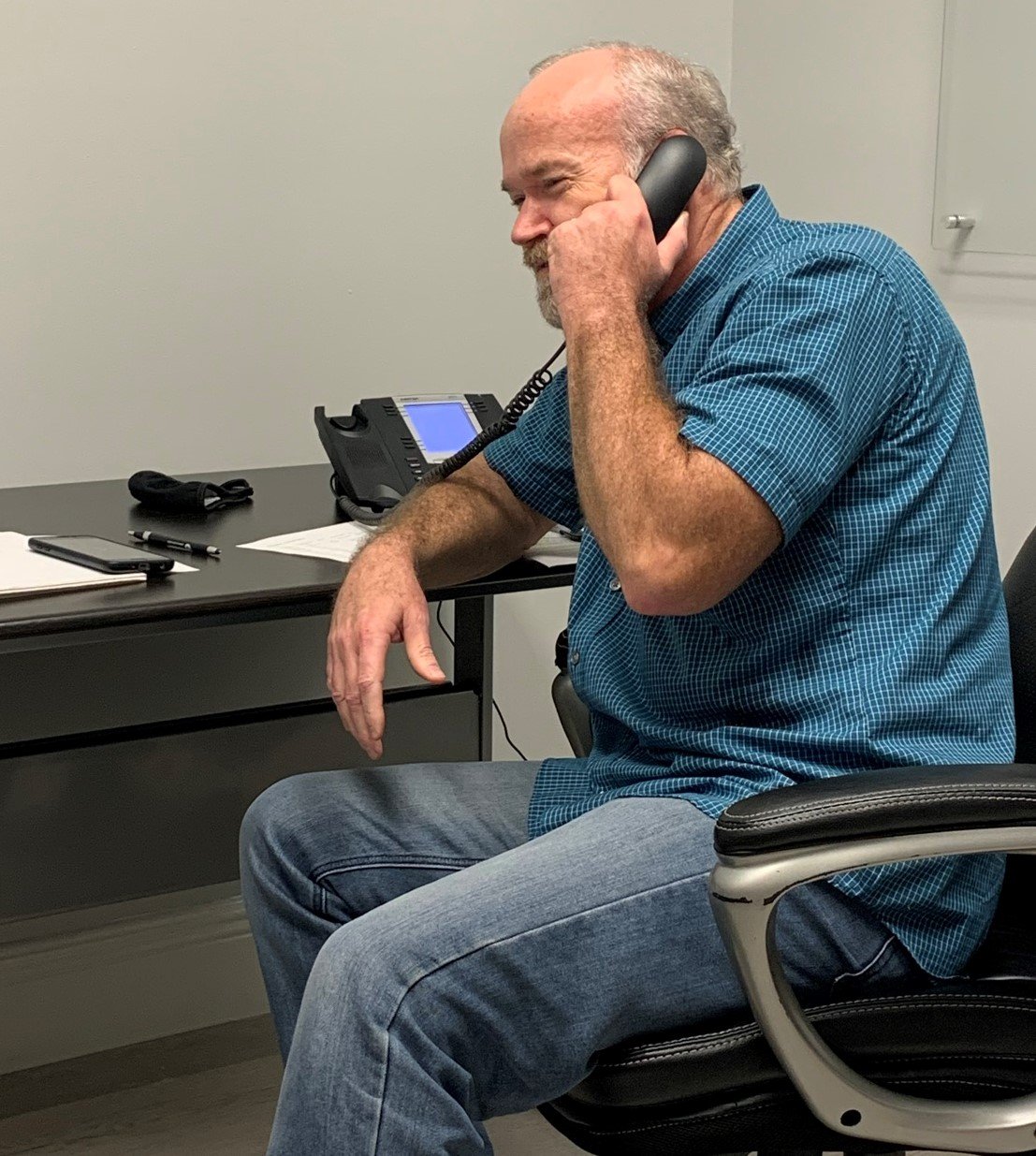 A man with a blond beard makes a phone call using a landline at a black, mostly empty desk.